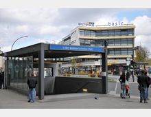 U-Bahnhof-Eingang Walther-Schreiber-Platz mit Glasüberdachung, im Hintergrund ein modernes Geschäftsgebäude mit der Beschriftung „Ärztehaus“ und „basic“. Fußgänger und Straßenverkehr an einer belebten Kreuzung.