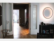 Moderner Wohnraum mit hellem Holzboden, weißen Wänden und einer großen, beleuchteten Kreis-Wandleuchte. Im Hintergrund sind ein Schreibtischstuhl, eine Glastür mit Blick ins Freie und ein Bücherregal mit Dekoobjekten zu sehen.