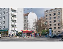 Straßenszene in einer urbanen Umgebung mit mehrstöckigen Wohnhäusern. Im Vordergrund ein kleiner Kiosk mit rotem Dach, Fahrradständern und Passanten, daneben eine Straßenkreuzung mit Ampel und parkenden Autos.