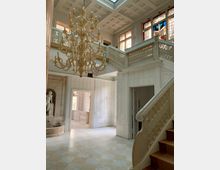 Großzügiger, historischer Innenraum mit einer weißen Kassettendecke, einem großen Kronleuchter, verzierten Balustraden und farbigen Buntglasfenstern. Rechts eine geschwungene Treppe, links ein Brunnen mit einer Statue in einer Wandnische.