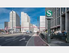 Straßenszene am Potsdamer Platz in Berlin mit modernen Hochhäusern im Hintergrund, einem S-Bahn-Schild im Vordergrund und rosa Rohren, die quer über die Straße verlaufen. Auf der rechten Seite befinden sich Geschäfte und Fußgänger, während Fahrräder an einem Geländer abgestellt sind.