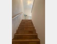 Eine Innentreppe mit braunen Fliesenstufen führt nach oben zu einem hellen Raum. Am oberen Ende der Treppe steht eine weiße Kommode mit dekorativen Objekten, darunter eine blaue Vase und ein Blumenstrauß.