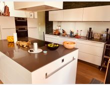 Moderne Küche mit weißen Einbaumöbeln, einer großen Kücheninsel mit dunkler Arbeitsplatte und integriertem Kochfeld. Im Hintergrund befindet sich eine Arbeitsfläche mit Haushaltsgeräten und grüner Fliesenrückwand, während Obstschalen und Deko-Elemente die Insel und die Arbeitsfläche schmücken.