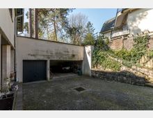 Eine gepflasterte Einfahrt mit einer Doppelgarage, von der eine Hälfte geöffnet ist und ein geparktes Auto sowie Fahrräder sichtbar sind. Im Hintergrund befinden sich ein mit Efeu bewachsener Hang, eine Steintreppe mit Geländer und einzelne Bäume.