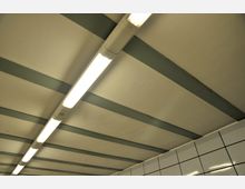 Decke eines Innenraums mit sichtbaren Metallträgern und langen, leuchtenden Deckenleuchten. Im unteren Bildbereich ist eine mit weißen Fliesen verkleidete Wand zu erkennen.