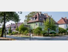 Mehrfamilienhäuser mit roten Ziegeldächern, umgeben von grünen Gärten und Bäumen, entlang einer ruhigen Straße. Ein Straßenschild mit der Aufschrift „Hellweg“ ist im Vordergrund sichtbar.