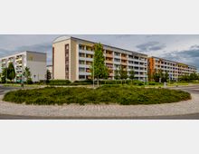 Mehrstöckige Wohngebäude mit Balkonen und farblich abgesetzten Fassaden, umgeben von kleineren Bäumen und gepflegten Grünflächen. Im Vordergrund ein runder, bepflanzter Verkehrskreisel mit gepflastertem Rand.