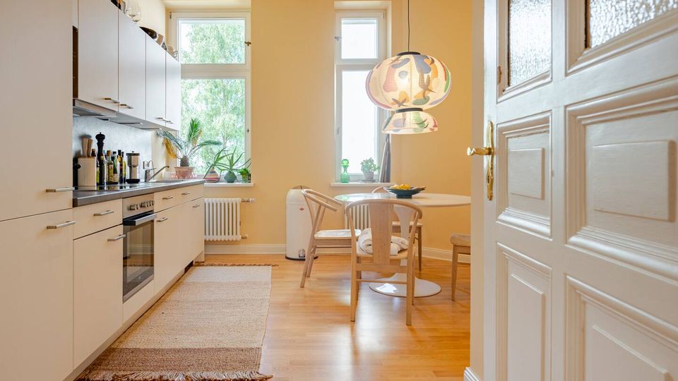Helle Wohnküche mit weißen Einbauschränken entlang einer Wand, Holzfußboden und kleinem Essbereich mit rundem Tisch und Stühlen. Zwei hohe Fenster bringen Tageslicht hinein; über dem Tisch hängen gemusterte Papierlampen, daneben sind Pflanzen und eine weiße Tür im Vordergrund zu sehen.