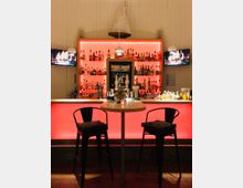 Barbereich mit beleuchtetem Tresen in Rot, darüber Regale mit großer Auswahl an alkoholischen Getränken. Zwei schwarze Barhocker und ein Stehtisch im Vordergrund, dekoriert mit einer kleinen Kerze; an den Wänden hängen zwei Bildschirme und ein Segelboot-Modell auf dem Regal.