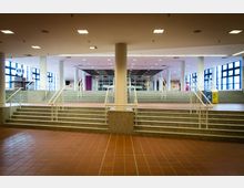 Weitläufiges Foyer eines öffentlichen Gebäudes mit symmetrischen Treppen aus Granit, die zu einer erhöhten Ebene führen. Große Fensterflächen lassen Tageslicht herein, und die Decke ist mit quadratischen Deckeneinbauleuchten versehen.