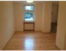 Schmaler, leerer Raum mit hellen Wänden und einem Boden aus hellem Holz. Ein großes Fenster mit Heizkörper darunter befindet sich an der Stirnseite, daneben führt eine kleine Treppe zu einer Tür.