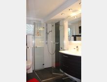 Modernes Badezimmer mit einer Dusche mit gläsernen Türen, einem dunklen Waschtisch mit integriertem Waschbecken und einem großen Spiegel. Die Wände sind weiß gefliest, und der Boden ist in dunklen Fliesen gehalten.