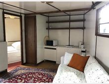 Wohnbereich in einem kleinen Raum mit weißen Wänden und Holzdetails. Zu sehen sind ein weißes Sofa mit Kissen, ein Teppich mit orientalischem Muster, ein kleiner Schrank mit Mikrowelle und Wasserkocher sowie Regalflächen an der Wand; links ist ein Durchgang zu einem Schlafzimmer sichtbar.