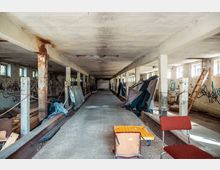 Verlassene Lagerhalle mit sichtbaren Betonstützen, beschädigter Decke und Wänden mit Graffiti. Im Raum befinden sich verstreut Möbel, Baumaterialien und ein improvisiertes Zelt; seitlich stehen kleine Fensterreihen, die schwaches Licht einfallen lassen.