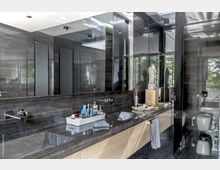 Modernes Badezimmer mit dunklen, glänzenden Marmorwänden und -oberflächen, einem breiten Waschbecken mit integriertem Wasserhahn, großem Wandspiegel und hellen Holzschränken. Im Hintergrund befinden sich eine Glasdusche und eine Toilette, während Dekorationsobjekte wie eine Statue und Raumduft auf der Ablage stehen.