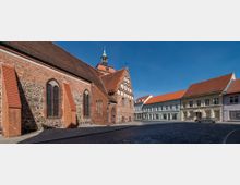 Historische Backsteinkirche mit angedeutetem Feldsteinfundament und spitzbogigen Fenstern, flankiert von Gebäuden mit hell verputzten Fassaden und roten Ziegeldächern entlang einer gepflasterten Straße unter blauem Himmel.