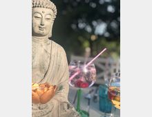 Steinstatue eines Buddhas mit einer goldenen Lotusblume, daneben stehen dekorative Gläser mit Getränken vor einem unscharfen, grünen Hintergrund im Freien.
