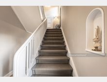 Innenbereich eines Treppenhauses mit einer mit Teppich belegten Treppe, weißen Wänden und einem weißen Geländer. Eine Nische in der Wand rechts enthält eine Hasenfigur, und oben an der Treppe befindet sich ein beleuchteter Flur.