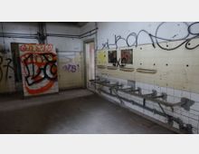 Ein verlassenes, heruntergekommenes Gebäude mit weißen Kacheln an den Wänden, die mit Graffiti bedeckt sind. An der rechten Wand sind beschädigte Halterungen und leere Ablageflächen zu sehen, während der Boden stark abgenutzt ist.