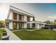Modernes, zweigeschossiges Wohnhaus mit flachem Pultdach, weiß-grauer Fassade und großen Fenstern mit Lamellenjalousien. Im Vordergrund liegen ein gepflegter Rasen, ein gepflasterter Weg und eine angrenzende Garage mit Einfahrt, eingefasst von einem dunklen Zaun in einer Wohnsiedlung.