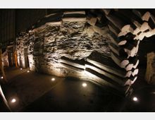 Eine Innenaufnahme einer felsigen Wandstruktur mit markanten Schichtungen, die von unten durch mehrere runde Bodenlichter in warmem Licht angestrahlt wird. Der Boden besteht aus dunklen Fliesen, und im Hintergrund ist ein Teil einer Holzstruktur erkennbar.