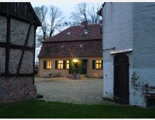 Ein gepflasterter Innenhof mit einem gelb gestrichenen Haus im Hintergrund, das grüne Fensterläden und ein rotes Ziegeldach hat. Rechts und links im Vordergrund stehen ältere Gebäude aus gemauertem und verputztem Stein sowie Fachwerk.