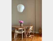 Ein minimalistischer Essbereich mit einem runden Holztisch und vier Stühlen, darunter ein Kinderstuhl. Auf dem Tisch steht eine Vase mit rosa Blumen. Eine weiße Papierlampe hängt von der Decke, und der Holzboden sowie ein brauner Tonkrug am Rand des Raumes sind sichtbar.