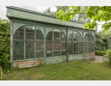 Grüne historische Gartenlaube mit verzierten Glasfenstern in Arkadenform, auf einem Backsteinsockel stehend, umgeben von gepflegtem Grün und Sträuchern. Eine goldene Inschrift ziert den oberen Rahmen der Struktur.