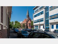 Straßenansicht mit geparkten Autos, einer Backsteinkirche mit spitzem Turm und roten Dachziegeln im Hintergrund sowie einem modernen weißen Gebäude mit blauen Fenstern rechts. Im Hintergrund sind Bäume und Wohnhäuser erkennbar.