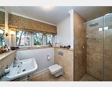 Modernes Badezimmer mit einer verglasten Dusche, einem Wand-WC und einem Waschbecken. Die Wände und der Boden sind mit beigem Naturstein verkleidet, ein großes Fenster mit gemustertem Rollo lässt Tageslicht herein.