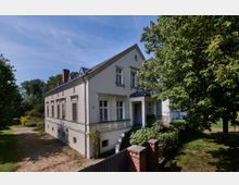 Einfamilienhaus im klassizistischen Stil mit heller Fassade, symmetrisch angeordneten Fenstern und einem kleinen Vordach über der Eingangstreppe, umgeben von Bäumen und einem Garten. Im Vordergrund ist ein Zaun mit einem gemauerten Torpfosten zu sehen.