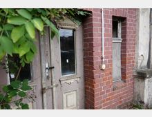 Eine alte Holztür mit abgeblätterter Farbe und Glasscheiben, neben einer Fassade aus rotem Backstein mit einem kleinen Fenster. Im Vordergrund hängen grüne Blätter von einer Pflanze herab.