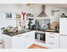 Kleine, moderne Küche mit weißem Eck-Unterschrank, Holzarbeitsplatte und einer Edelstahlspüle. Im Hintergrund sind Wanddekorationen, ein roter Hummer aus Plastik, gerahmte Bilder, Regale mit Küchenutensilien sowie ein Herd mit Dunstabzugshaube sichtbar.