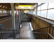 Bahnhof mit überdachten Bahnsteigen und einer Treppe, die zu einer unteren Ebene führt. Große Fensterreihen sorgen für Tageslicht, und Hinweisschilder mit gelbem Hintergrund geben Informationen zu Fahrtrichtungen und Zielen wie „U6 Jüdisches Museum“.