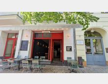 Außenansicht eines Cafés oder Bistros mit dem Namen "Melody Nelson", erkennbar an der Beschriftung über dem Eingang. Vor dem Eingang befinden sich grüne Metalltische und -stühle, daneben eine Schiefertafel mit Menüangeboten, während im Inneren eine Bar und Sitzgelegenheiten sichtbar sind.