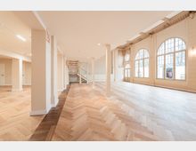 Großer, leerer Innenraum mit hellem Fischgrätparkett, vielen weißen Säulen und offener Treppe im Hintergrund. An der rechten Wand befinden sich hohe Rundbogenfenster und dekorativer Stuck unter einer hohen Decke.