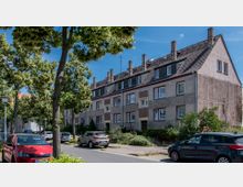 Reihenhäuser mit verputzten Fassaden, mehreren Fenstern und Dachgauben in einer ruhigen Wohnstraße, umgeben von Bäumen. Geparkte Autos stehen entlang der Straße, und der Gehweg ist von grünen Sträuchern gesäumt.