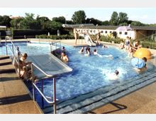 Außenbereich eines Schwimmbads mit mehreren Personen, darunter Kinder und Erwachsene, die im flachen Becken schwimmen oder am Rand entspannen. Zu sehen sind kleine Rutschen, ein Geländer, ein gepflasterter Bereich sowie eine Begrünung und Gebäude im Hintergrund.