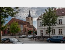 Straßenszene mit einer historischen Backsteinkirche auf der linken Seite, erkennbar an ihrem spitzen Turm und Rundbogenfenstern, sowie einem weißen Gebäude mit Apotheke rechts. Davor stehen geparkte Autos, Straßenlaternen und einige Bäume.
