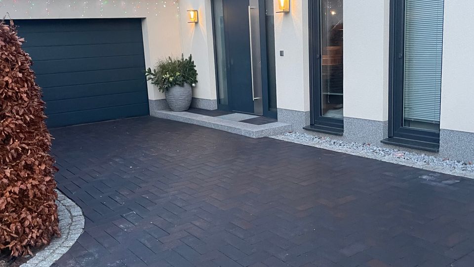 Moderne Hausfassade mit heller Putzoberfläche und großen Fenstern, daneben eine integrierte Garage mit dunkelgrauem Tor. Der Eingangsbereich ist mit einer Tür in anthrazitfarbenem Rahmen und Außenbeleuchtung gestaltet. Davor liegt eine gepflasterte Einfahrt.