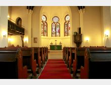 Innenraum einer Kirche mit Holzbänken, einem roten Teppich im Mittelgang und einem Altar mit grünem Altartuch. Im Hintergrund sind bunte Glasfenster und eine erhöhte Kanzel an der rechten Wand sichtbar.
