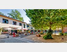 Ein ruhiger Innenhof mit Bäumen und Cafétischen, umgeben von niedrigen, hellen Gebäuden mit großen Fenstern und einzelnen Geschäften, darunter ein Theater. Holzbänke, Sonnenschirme und ein gepflasterter Weg prägen den Außenbereich.