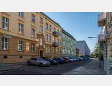 Straßenszene mit mehrstöckigen Wohnhäusern in gelber und grüner Farbe im klassischen Stil mit Fenstergesimsen und Balkonen. Geparkte Autos säumen den Straßenrand, und ein 30-km/h-Verkehrsschild ist sichtbar; der Himmel ist klar und die Straße ruhig.