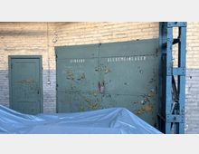 Außenwand eines Industriegebäudes aus weiß gestrichenem Ziegel mit einer kleinen Tür und einem großen, abblätternden grünen Doppeltor mit der Aufschrift „Eingang“ und „Allgemeinlager“. Rechts steht eine blaue Stahlkonstruktion, im Vordergrund liegt eine graue Abdeckplane.