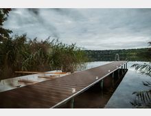 Ein ruhiger See mit einem schmalen Holzsteg, der in das Wasser führt. Neben dem Steg liegt ein Ruderboot, umgeben von Schilf, während der Himmel bewölkt ist.