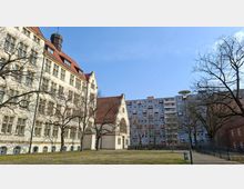 Innenhof mit Rasenfläche und Gehweg, umgeben von einem historischen hellen Gebäude mit steilem roten Ziegeldach, vielen Fenstern und einem kleinen Turm sowie einem hohen modernen Wohnblock im Hintergrund. Kahle Bäume, Laternen und Fahrradständer stehen am Rand des Platzes.