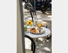 Ein runder Marmortisch auf einem Bürgersteig mit einer Mahlzeit bestehend aus einem Toast-Sandwich, einer Essiggurke und einer kleinen Soßenschale auf einem silbernen Tablett. Daneben stehen ein Glas Wasser, eine Karaffe, ein goldfarbenes Trinkglas, ein Glas mit einer Sonnenblume und ein grünes Getränk. Im Hintergrund sind gepflasterter Boden, Fahrrad und parkende Fahrzeuge sichtbar.