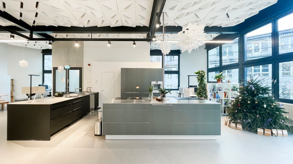 Großer, heller Innenraum mit moderner Kücheninsel und Arbeitszeilen, hohen Decken mit geometrischen Akustikpaneelen und schwarzen Deckenlampen. Rechts sorgen bodentiefe Fensterfront, Regalbereich mit Pflanzen und ein dekorierter Weihnachtsbaum für Kontext in einem offenen Büro- oder Gemeinschaftsbereich.