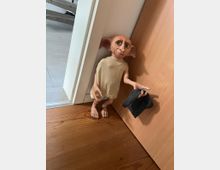 Eine Figur eines Hauselfen aus einem Film, die an einer Holztür in einem Innenraum lehnt. Der Boden besteht aus Holz, und im Hintergrund sind helle Fliesen sowie eine Treppenstufe zu sehen.