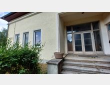 Eingangsbereich eines einfachen Gebäudes mit gelber Putzfassade, drei Fenstern auf der linken Seite und einer verglasten Doppeltür. Vor dem Eingang befindet sich eine kleine Treppe mit einem Blumentopf auf dem Geländer, daneben ist eine grüne Hecke sichtbar.